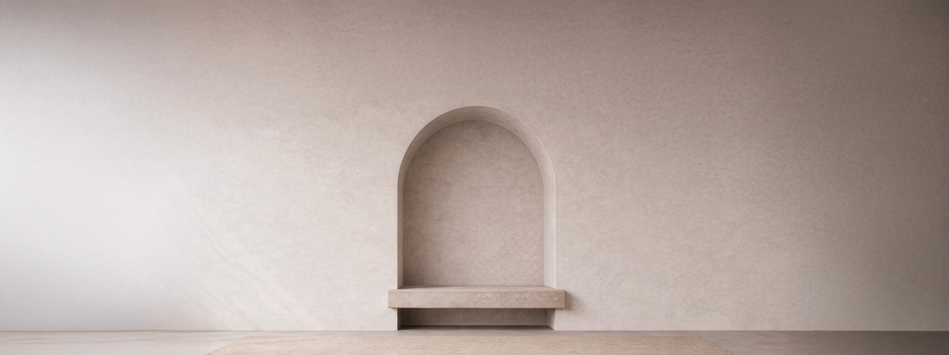 Empty arched wall niche in warm off-white plaster with a stone bench shelf, softly lit above a woven neutral floor.